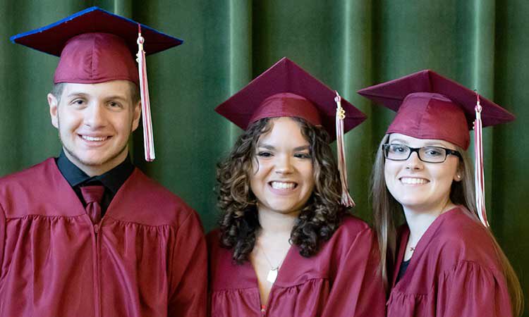 Smiling graduating students, one male and 2 females, wearing cap and gown.