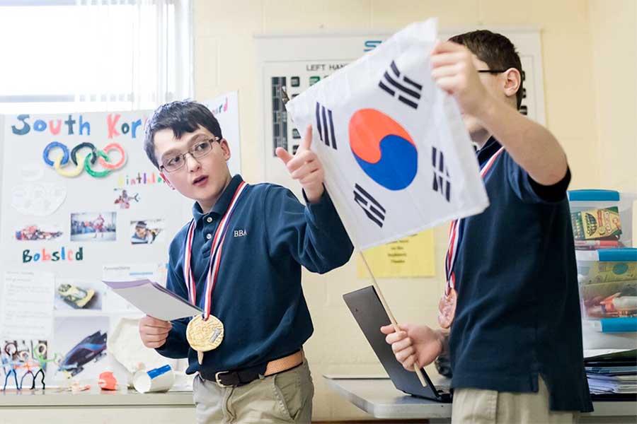 Two students holding the South Korean flag up; one student has a gold medal; posters with information about South Korea are hanging on the wall.