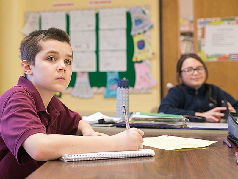 Male student sitting at desk with notebook, holding pen in his hand watching the teacher.