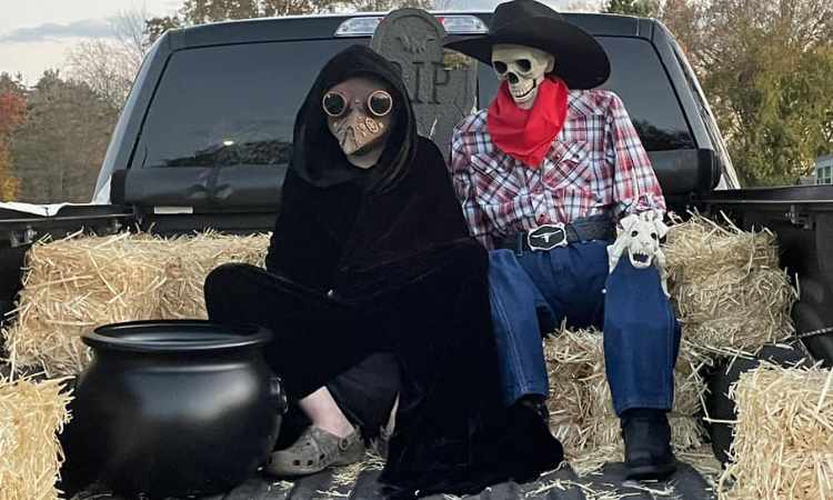 2 skeletons dressed as Death and Cowboy sitting on hay bales in back of pickup truck.
