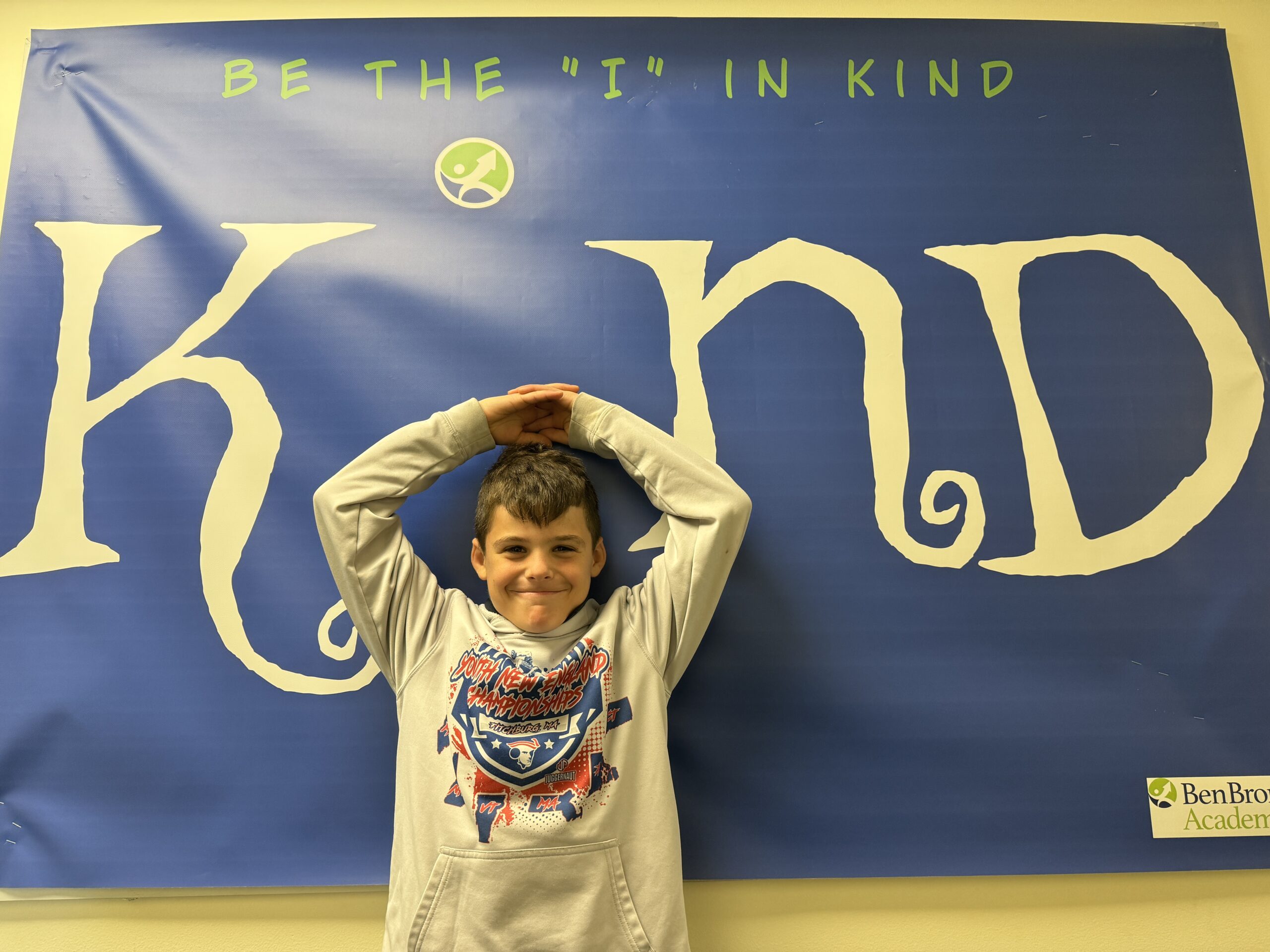 IMG_4917 Smiling male students holding his arms above his head, standing in front of banner replacing the letter "I" in the word "Kind."