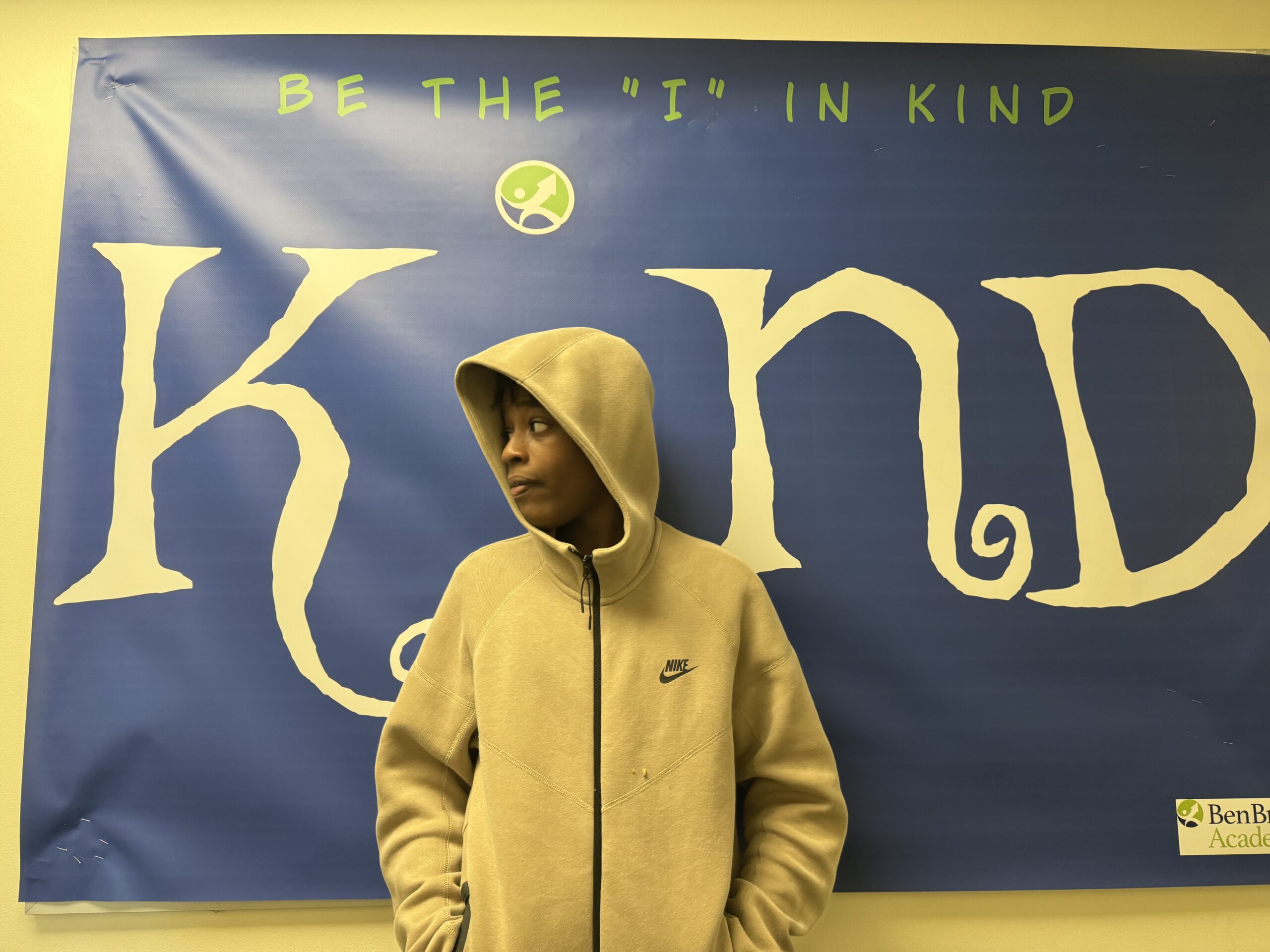 IMG_4923 Male student wearing a hooded sweatshirt, standing in front of banner replacing the letter "I" in the word "Kind."